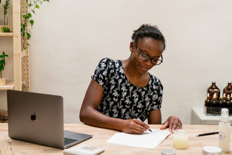 Woman writing at desk with laptop and jars.