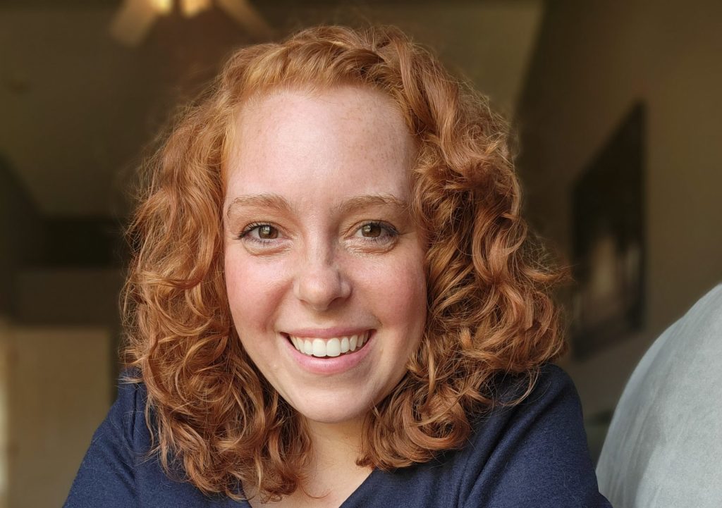 Smiling person with curly red hair indoors.